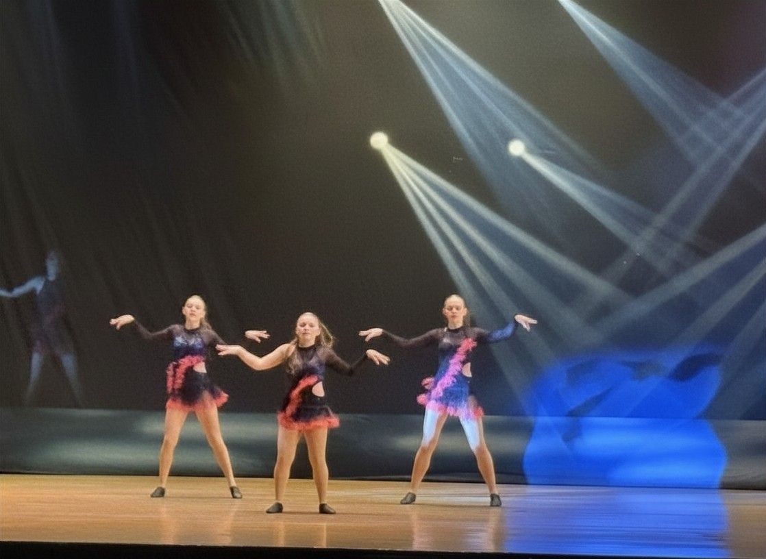 Three dancers in black and pink outfits perform on a stage under bright spotlights.