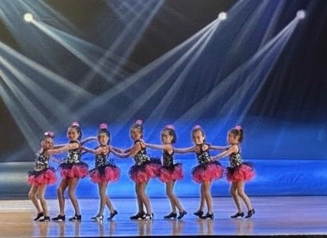 Six dancers in pink costumes perform on a lit stage under blue spotlights.