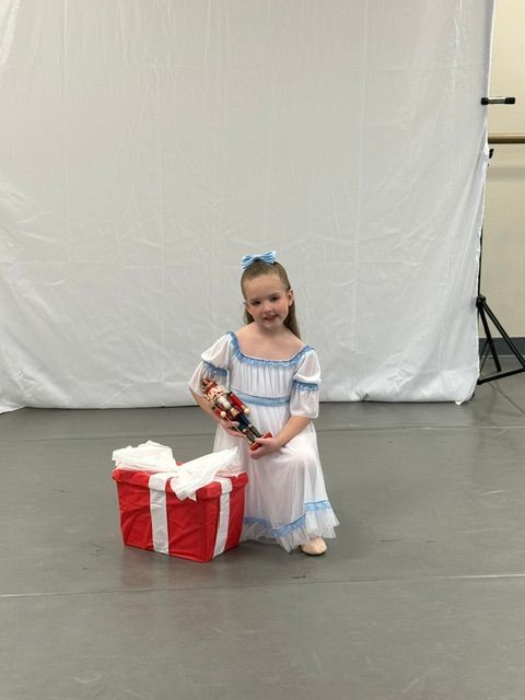Child kneeling beside a red gift box in a white dress, holding a wrapped present on a plain backdrop.
