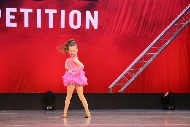 Young dancer in a pink costume performing on a stage with a red backdrop and ladder