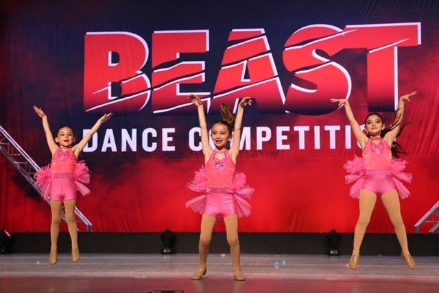 Three dancers in pink costumes performing onstage at a BEAST dance competition