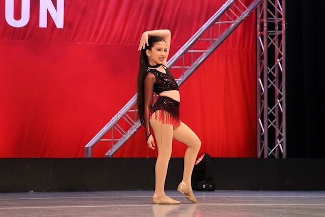 Young dancer posing on stage in black and red costume against a red backdrop