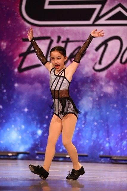 Young dancer in a black-and-silver costume performs on stage with arms raised
