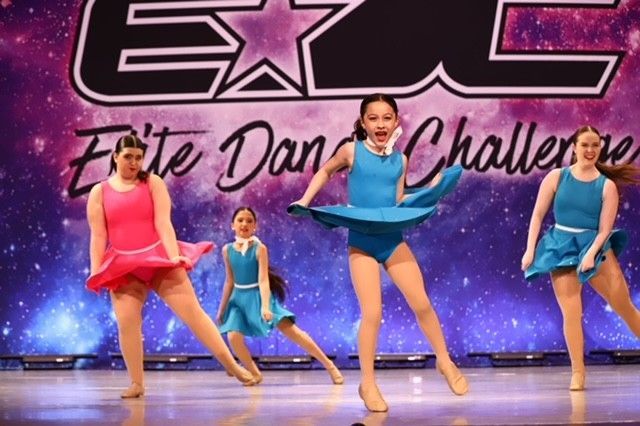 Four dancers in colorful costumes performing a high-kick routine on a stage backdrop