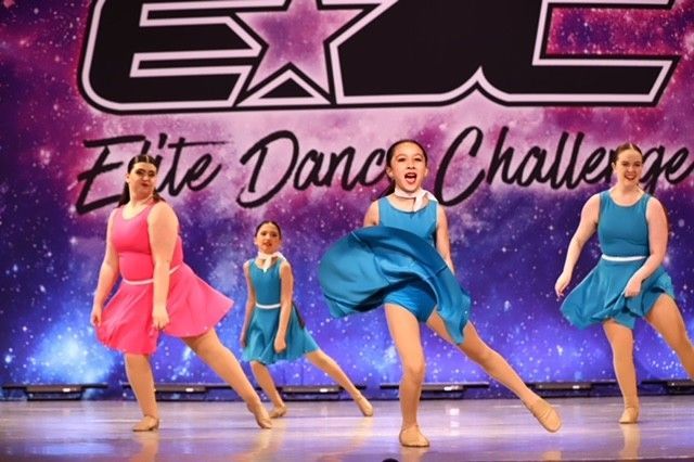 Four dancers in colorful dresses leap on a stage with an “Elite Dance Challenge” backdrop