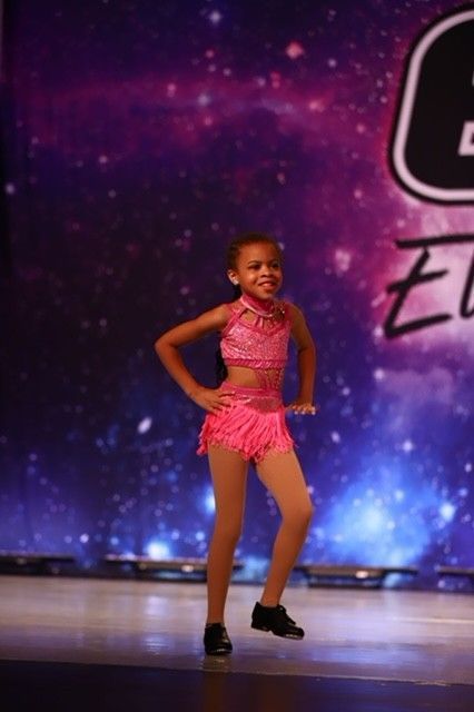 Young dancer in a pink costume poses on stage against a purple starry backdrop.