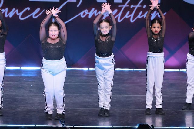 Four dancers onstage in black tops and white pants, arms raised during a performance.