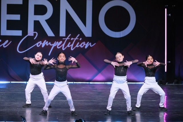 Four dancers in black tops and white pants perform on a lit stage with a “Competition” backdrop