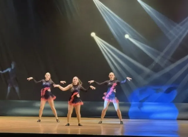 Three dancers in black and pink costumes perform on a stage with dramatic blue and white spotlights.