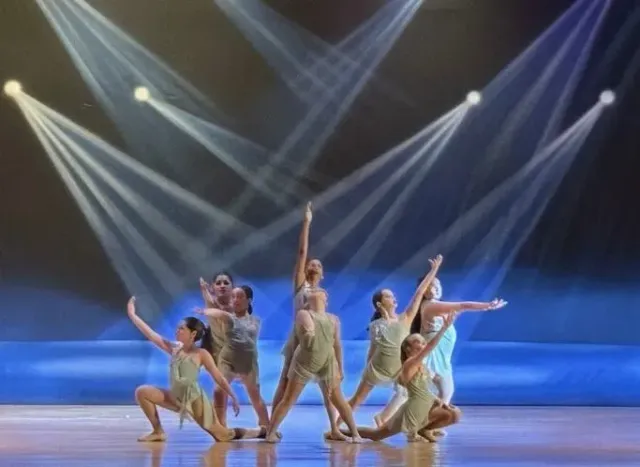 Dancers in beige costumes pose under bright stage spotlights on a blue-lit performance stage
