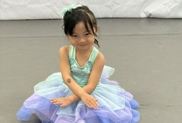Young child in a pastel dress sitting cross-legged on a gray floor, smiling softly.