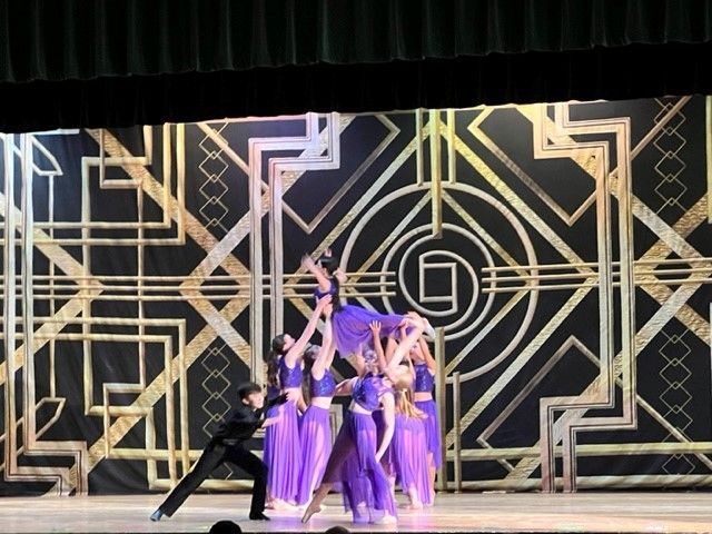 Dancers in purple and gold costumes performing on a stage with art deco backdrop. A group lifts a dancer.
