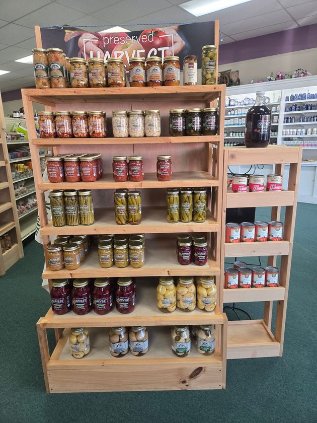 A wooden retail display stand stocked with various jars of preserved foods, pickles, and jams in a store setting.