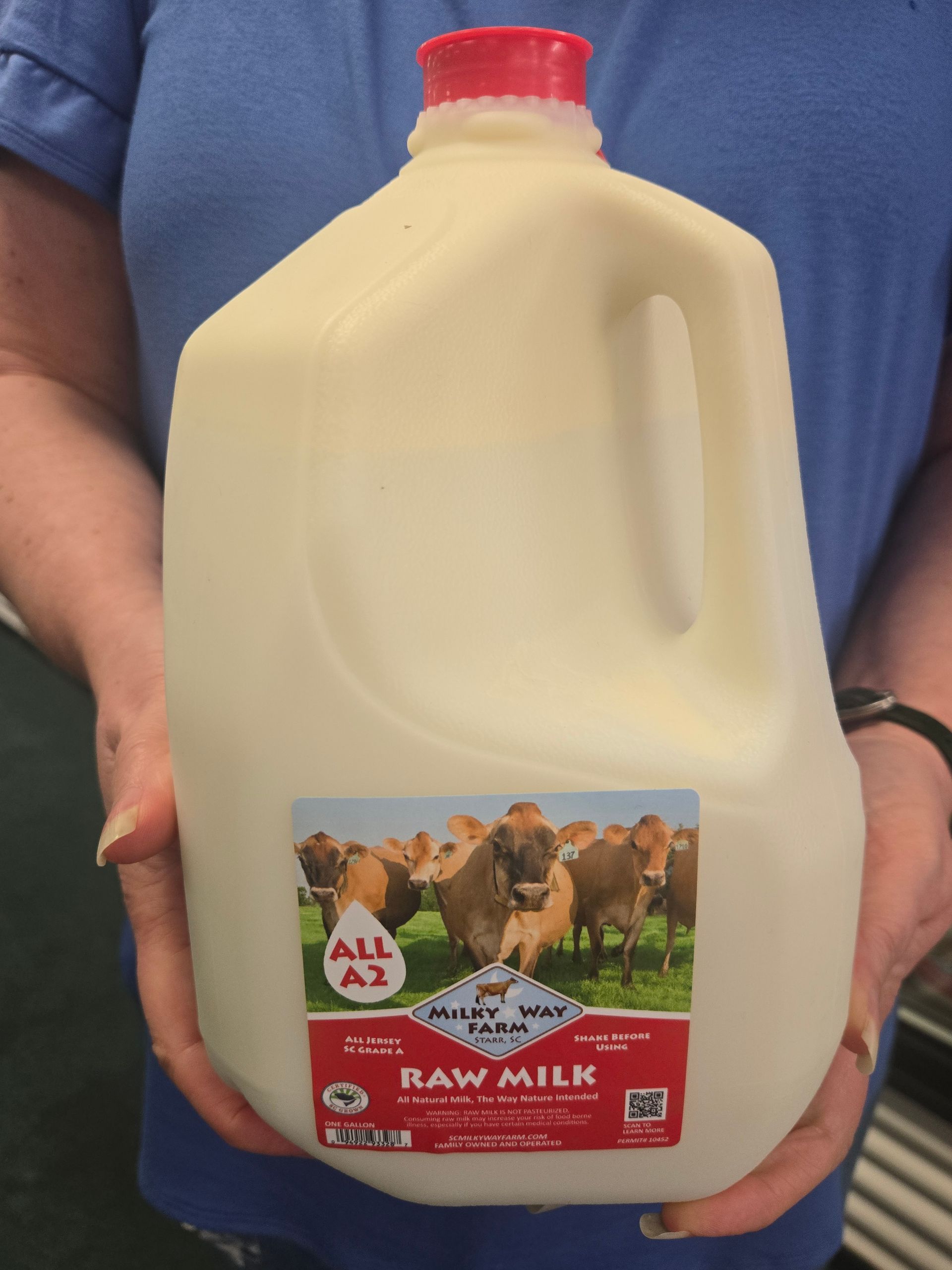 Hands holding a gallon jug of Milky Way Farm raw milk with a red cap and a label featuring cows.