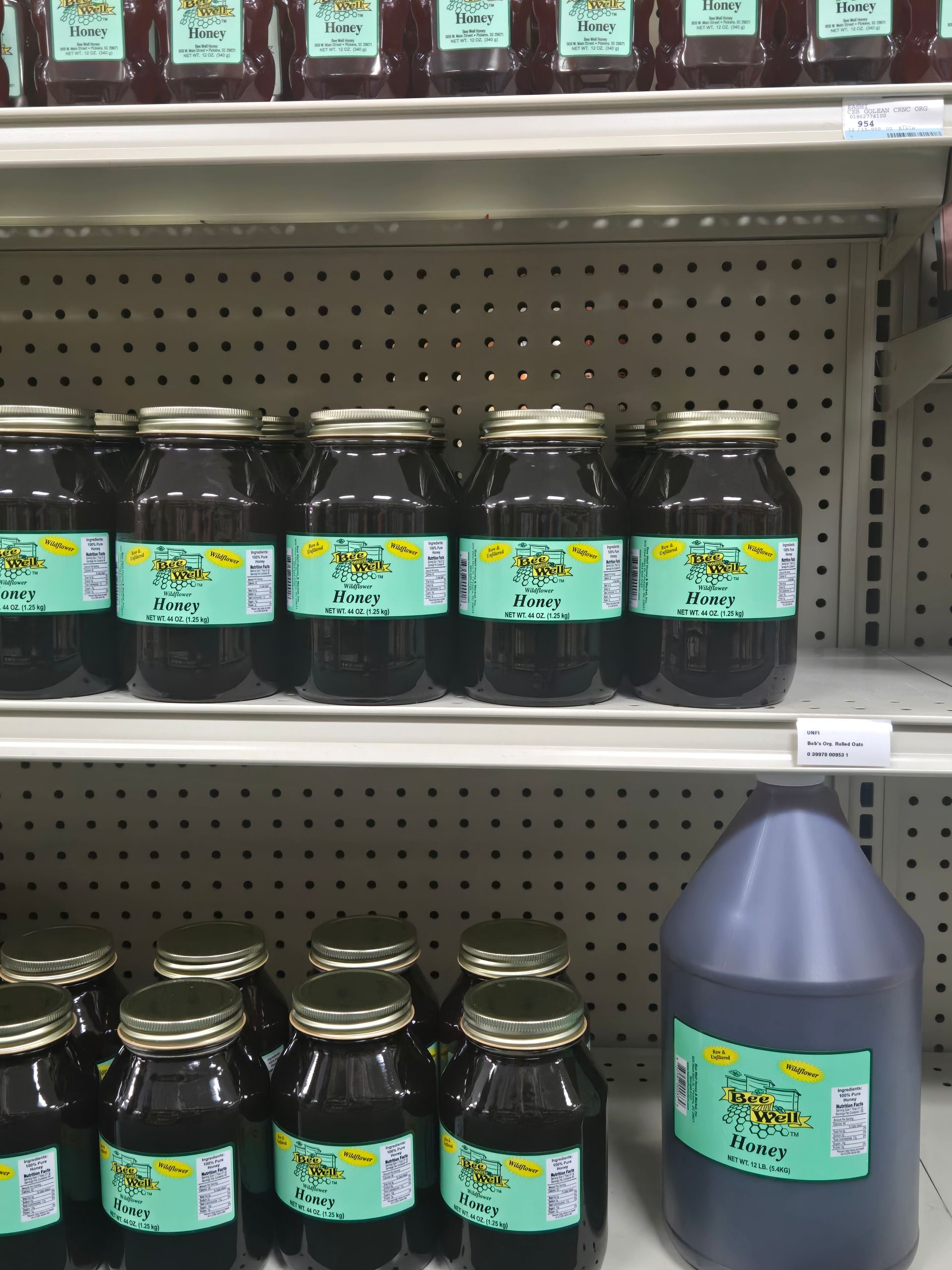 Shelves in a grocery store stocked with multiple jars and a large plastic jug of dark honey, all with light green labels.