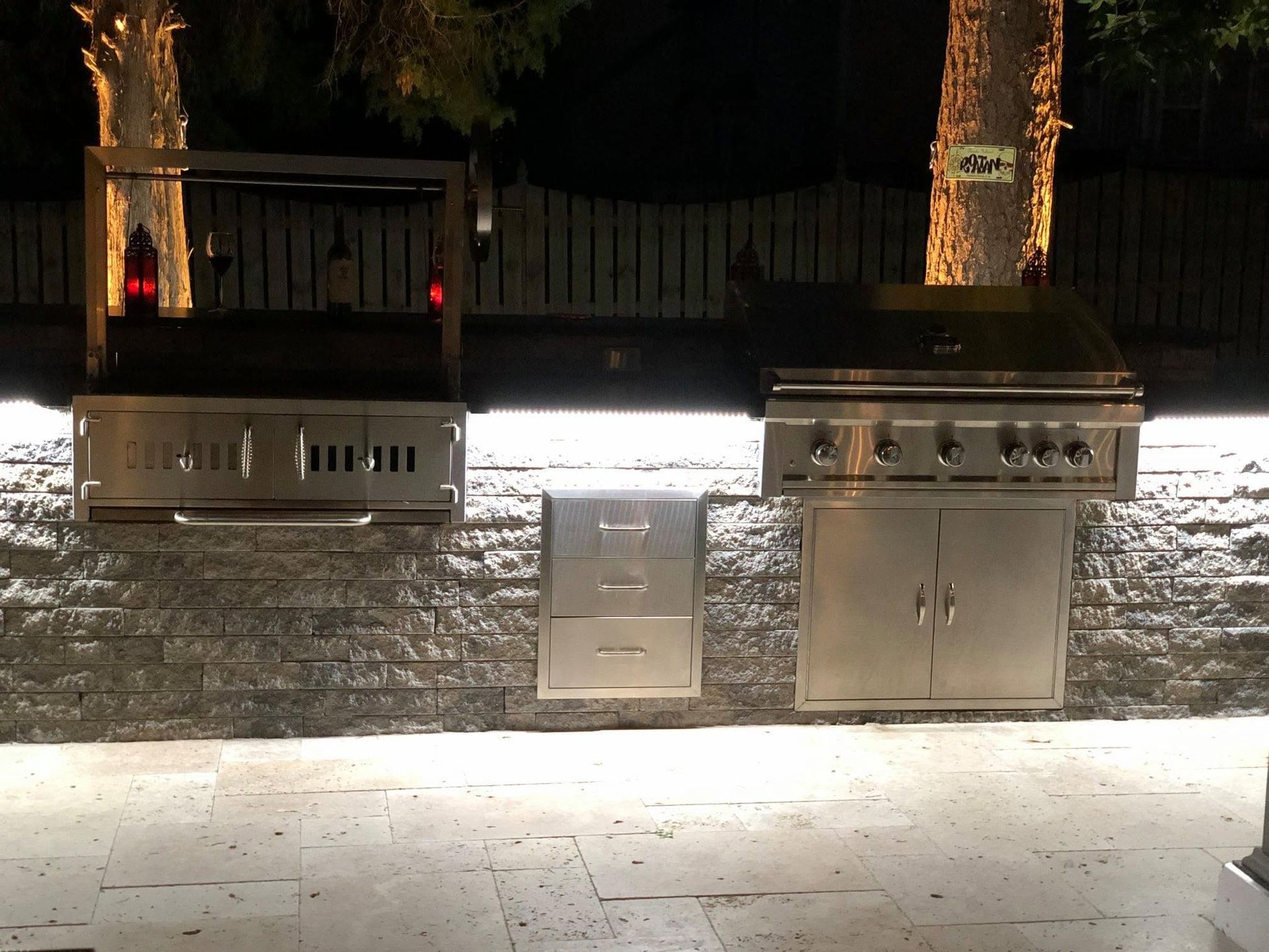 Outdoor stone wall with two stainless steel grills and a utility cabinet at night