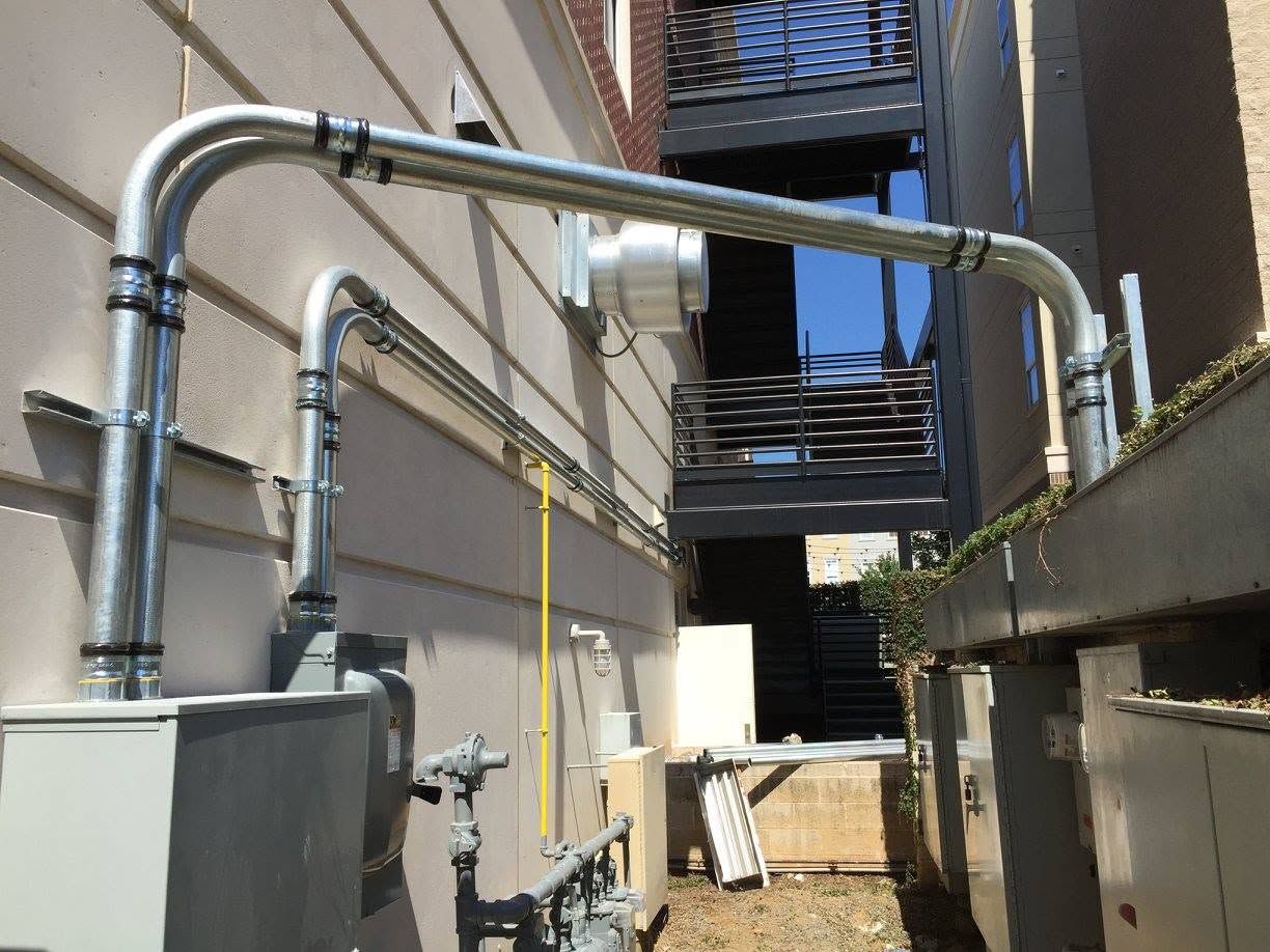 Narrow alley with large silver pipes running between buildings and a utility unit on the left.