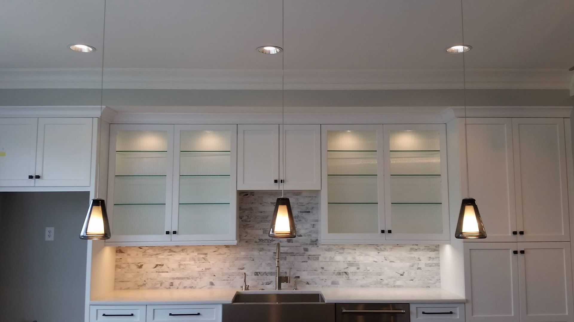 Modern white kitchen with pendant lights, glass-front cabinets, tiled backsplash, and built-in appliances