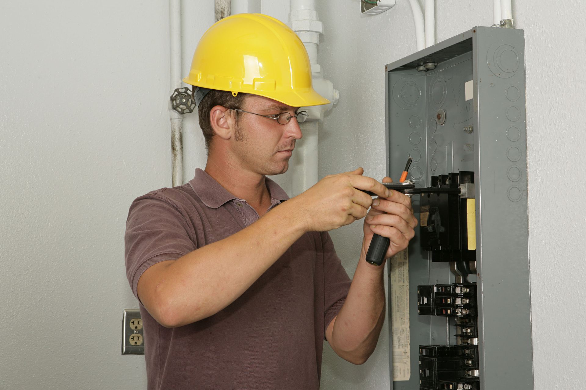 Electrician in yellow hard hat works on an open electrical panel with a screwdriver.