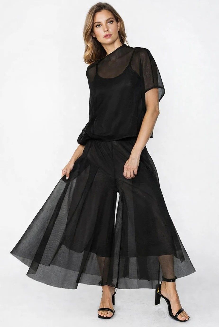 Woman in black sheer jumpsuit and sandals, standing, looking to the side.