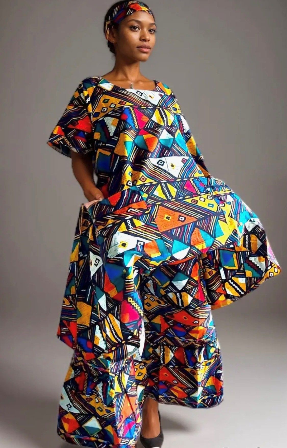 Woman in a colorful print dress and headscarf, hands in pockets, posing.