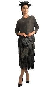 Woman in a dark, fringed dress and hat, holding a purse and wearing heels.