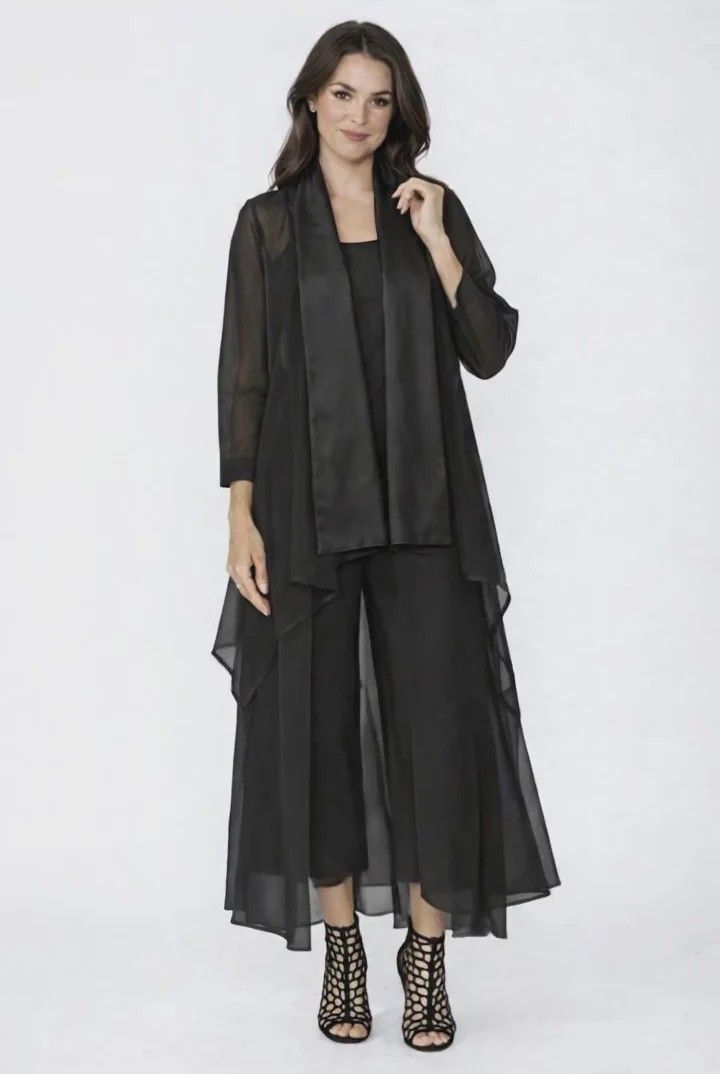 Woman in a black dress pantsuit and sheer duster, standing against a white backdrop.