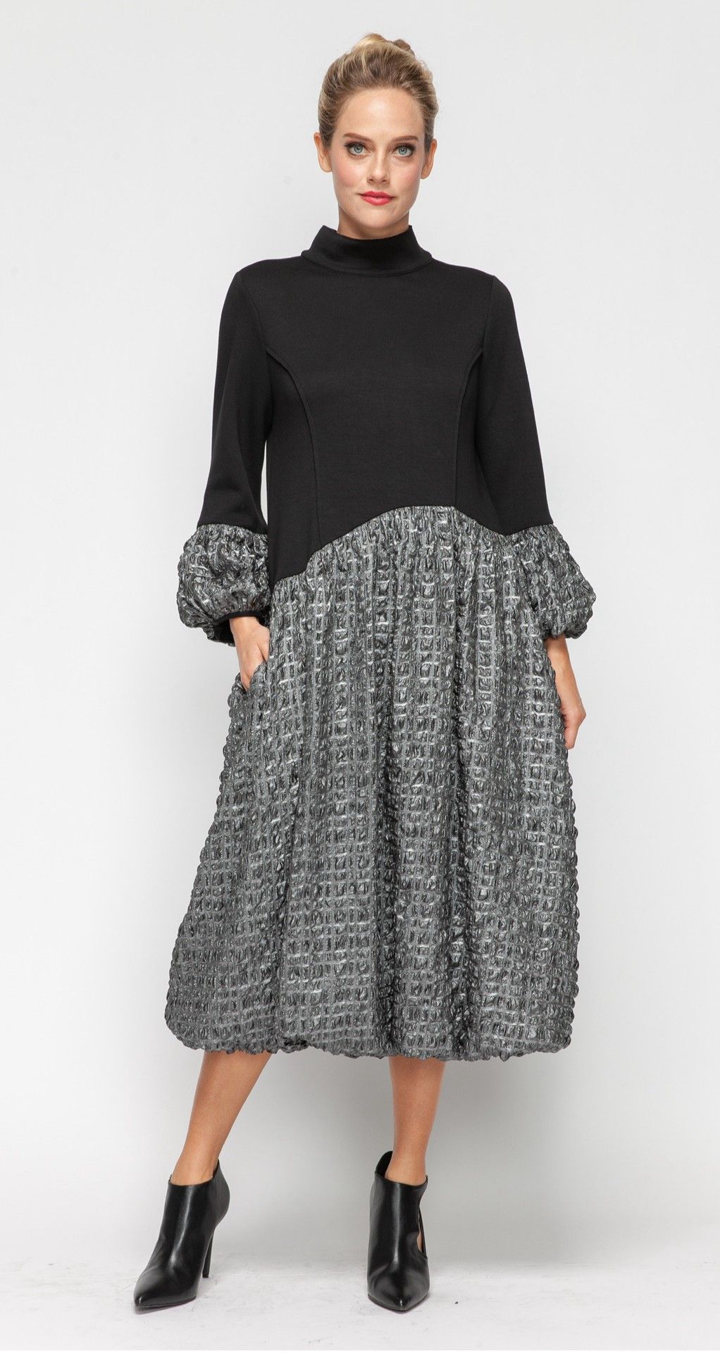 Woman in black and patterned dress with a high collar and ankle boots, posing in front of a white backdrop.