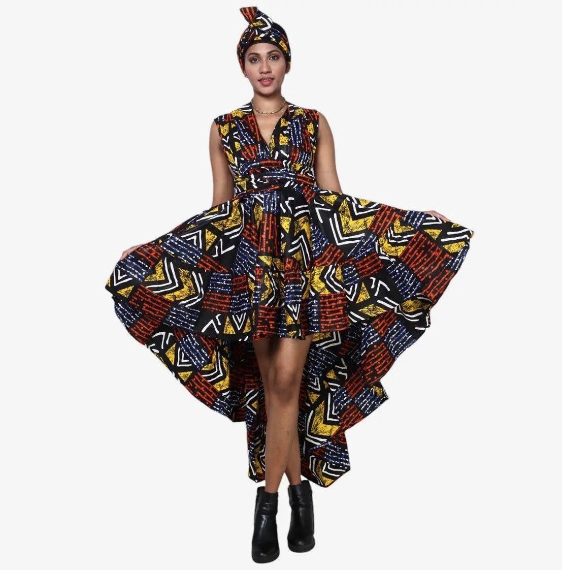 Woman in patterned high-low dress and headwrap, holding dress out, wearing black boots.