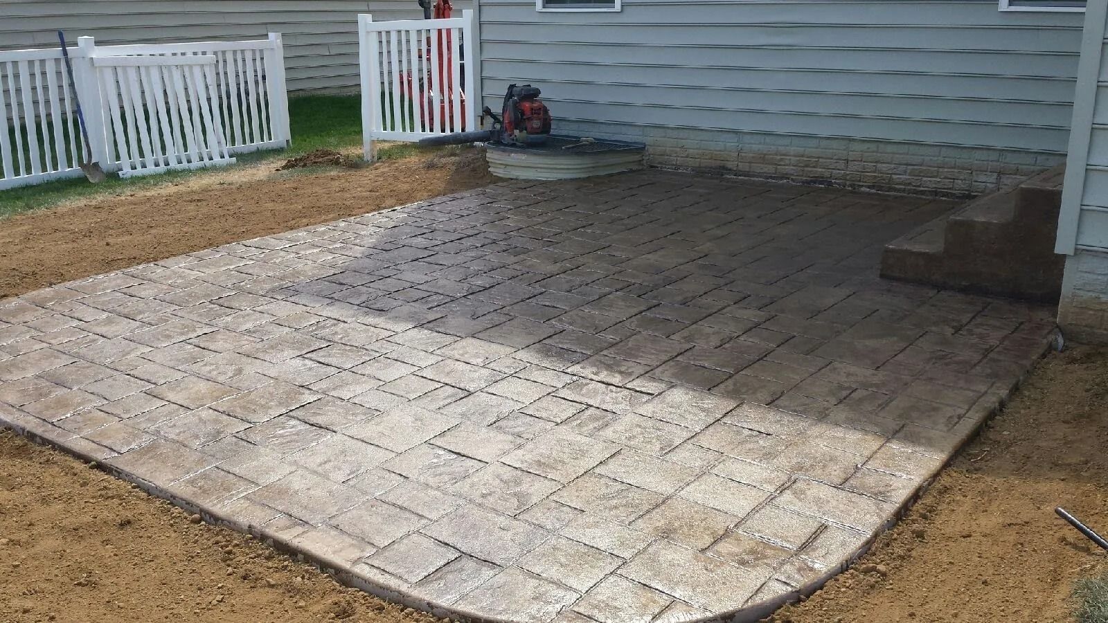 Brick patio next to a house with white fencing and dirt surrounding the patio.