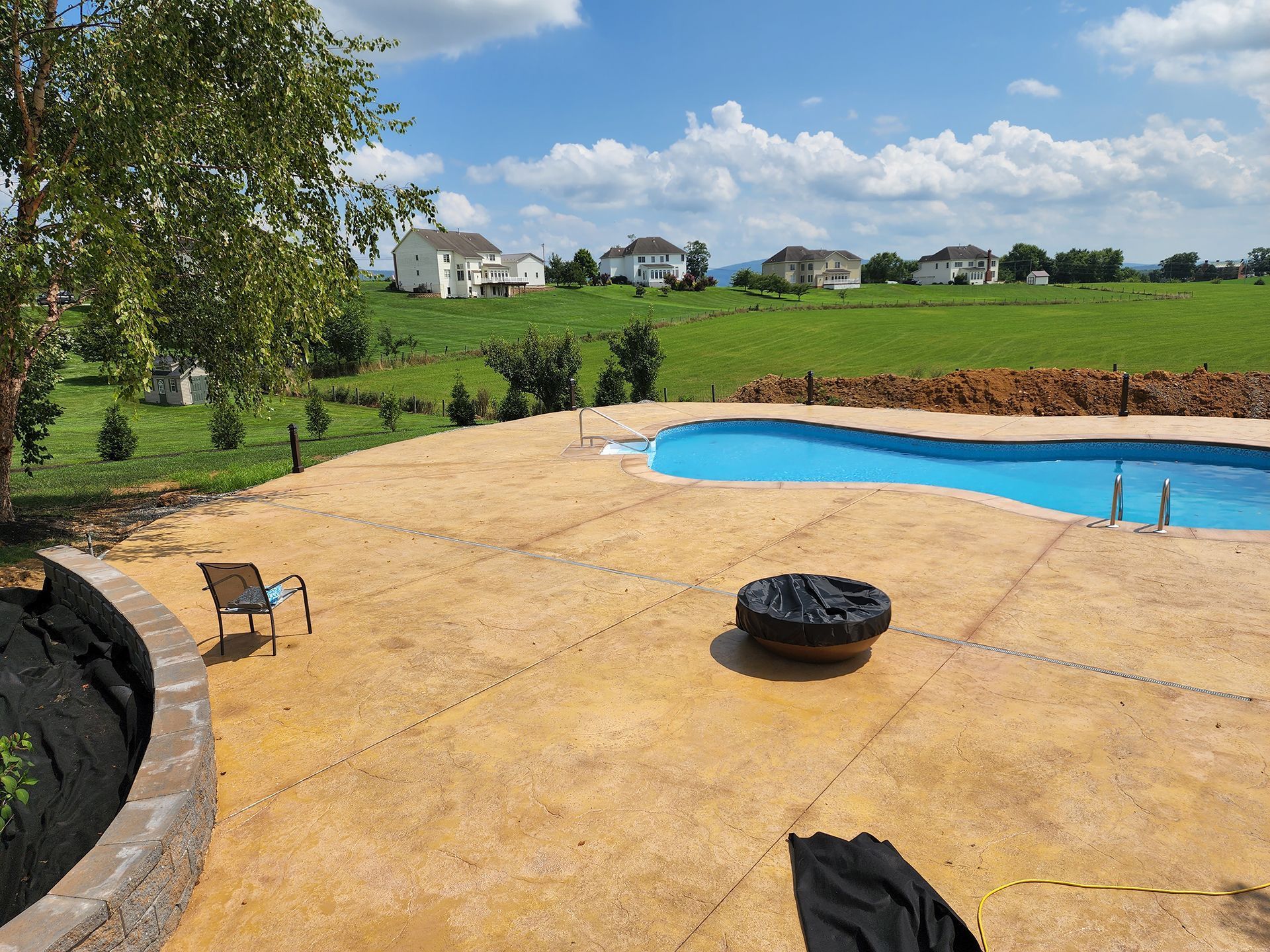 Backyard with pool, patio, fire pit, and view of houses and green field on a sunny day.