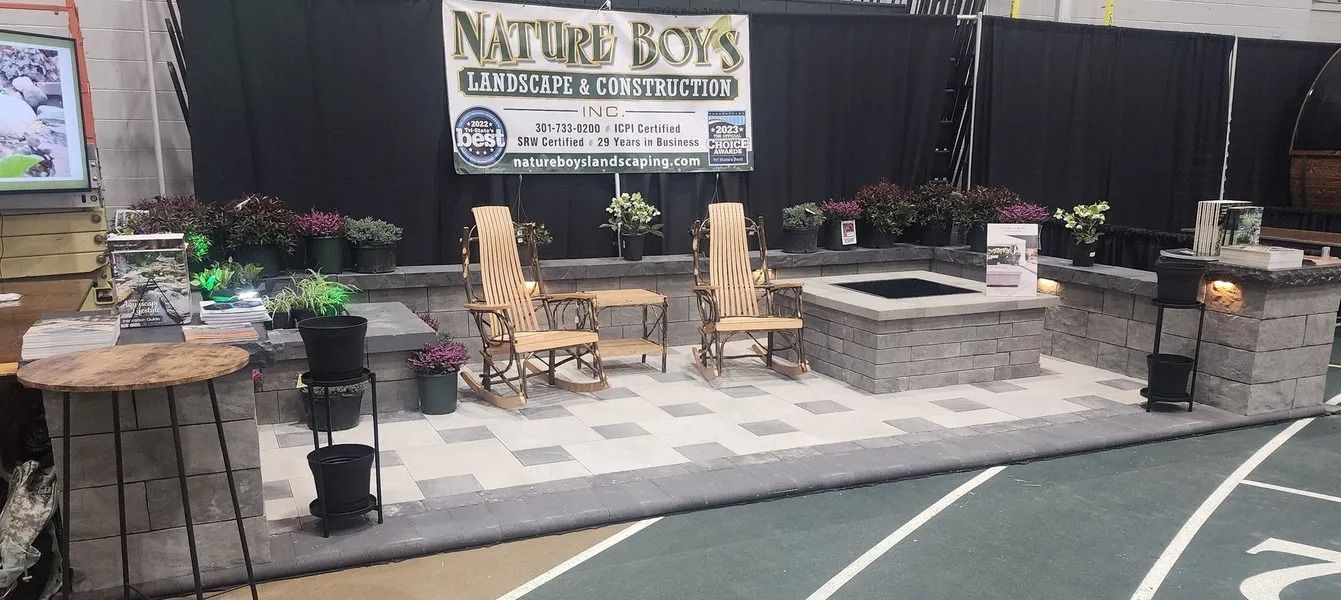 A trade show booth with landscaping display, including chairs, a fire pit, and potted plants. 