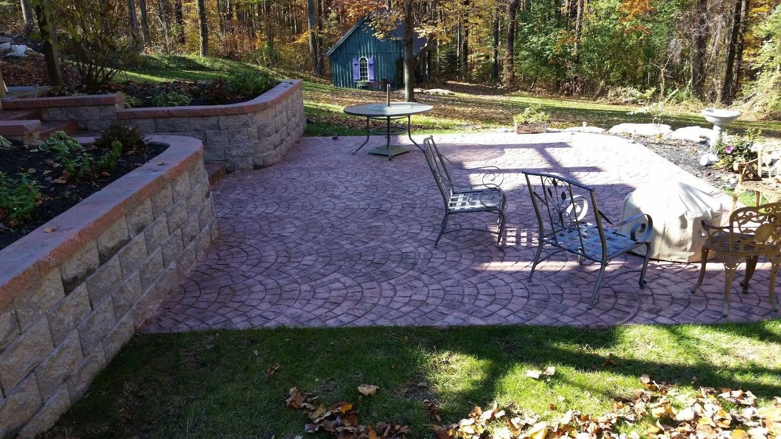 Stone patio with iron furniture, retaining wall, garden, and shed in a wooded yard.