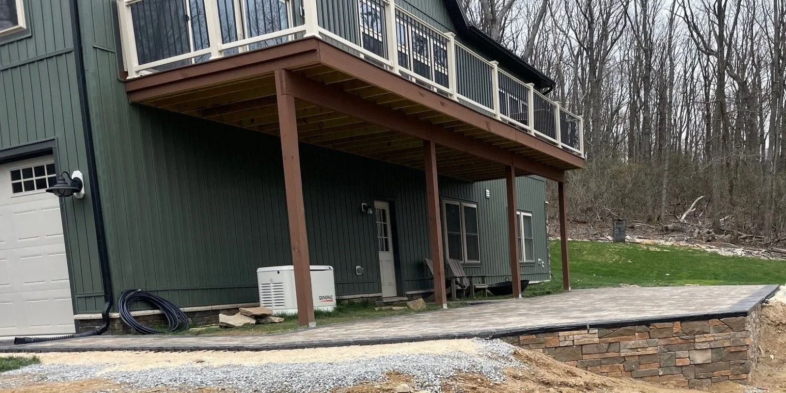 A house with a second-story deck, a stone patio, and a grassy yard, with a wooded area in the background.