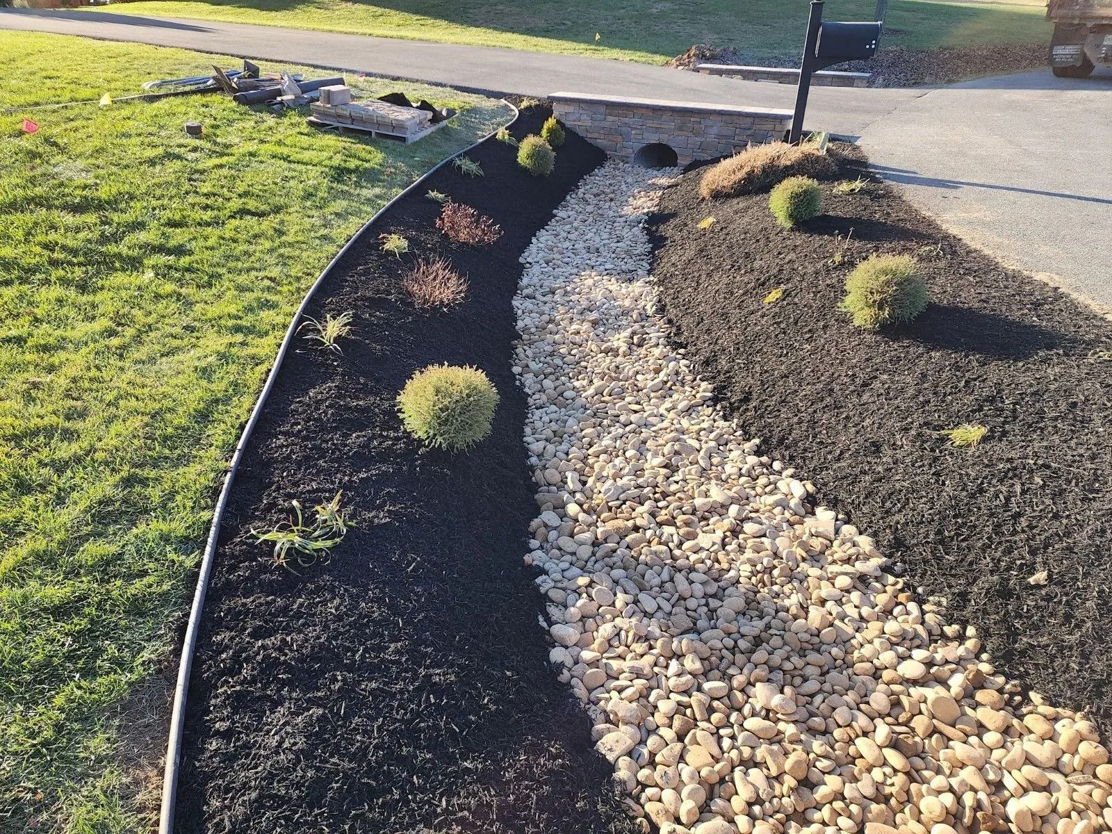 Landscaped bed with black mulch, tan rocks, shrubs, and a metal border.