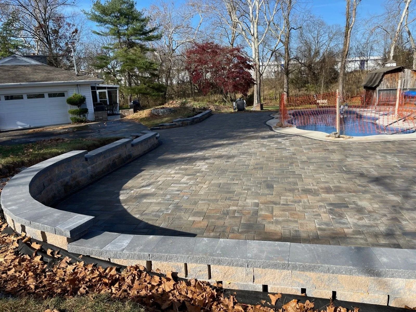 Brick patio and retaining wall surrounding a pool, next to a house and trees.