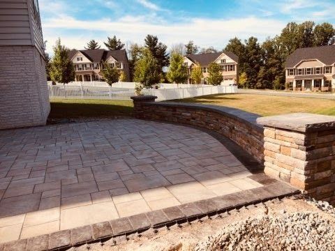 Paver patio with stone retaining wall, bordering a grassy area with houses and sky.