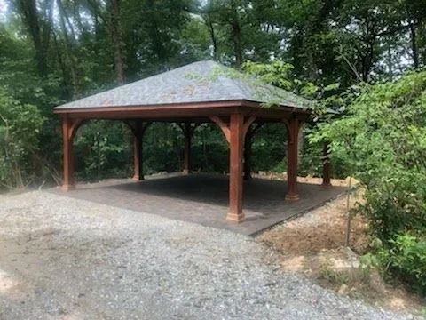 Wooden gazebo with a gray roof and brown posts, set on a stone foundation surrounded by trees and a gravel path.
