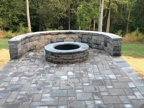 Stone fire pit with curved seating area on a paved patio, outdoors, surrounded by trees.