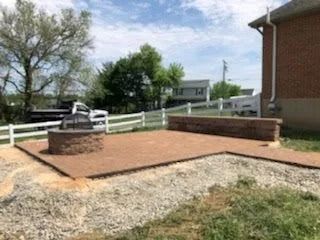 A brick patio with fire pit and retaining wall in a yard, with a white fence and house in the background.