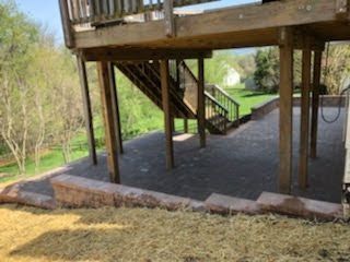 Wooden deck with stairs, supported by posts, over a stone patio, with a retaining wall.