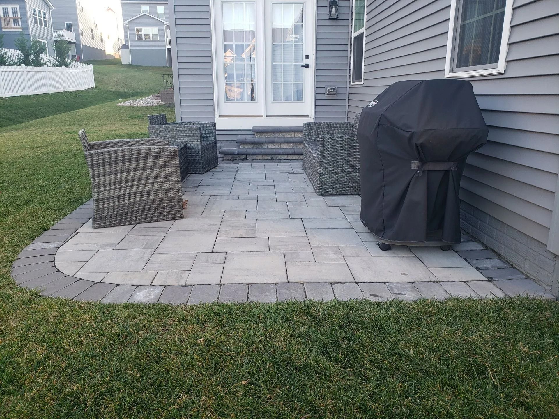 A paved patio with wicker chairs and a covered grill next to a gray house.