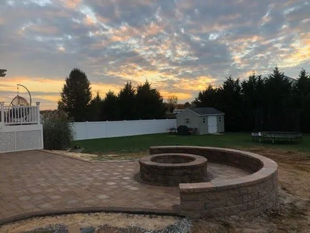 Backyard with a brick fire pit, patio, white fence, shed, and cloudy sunset.