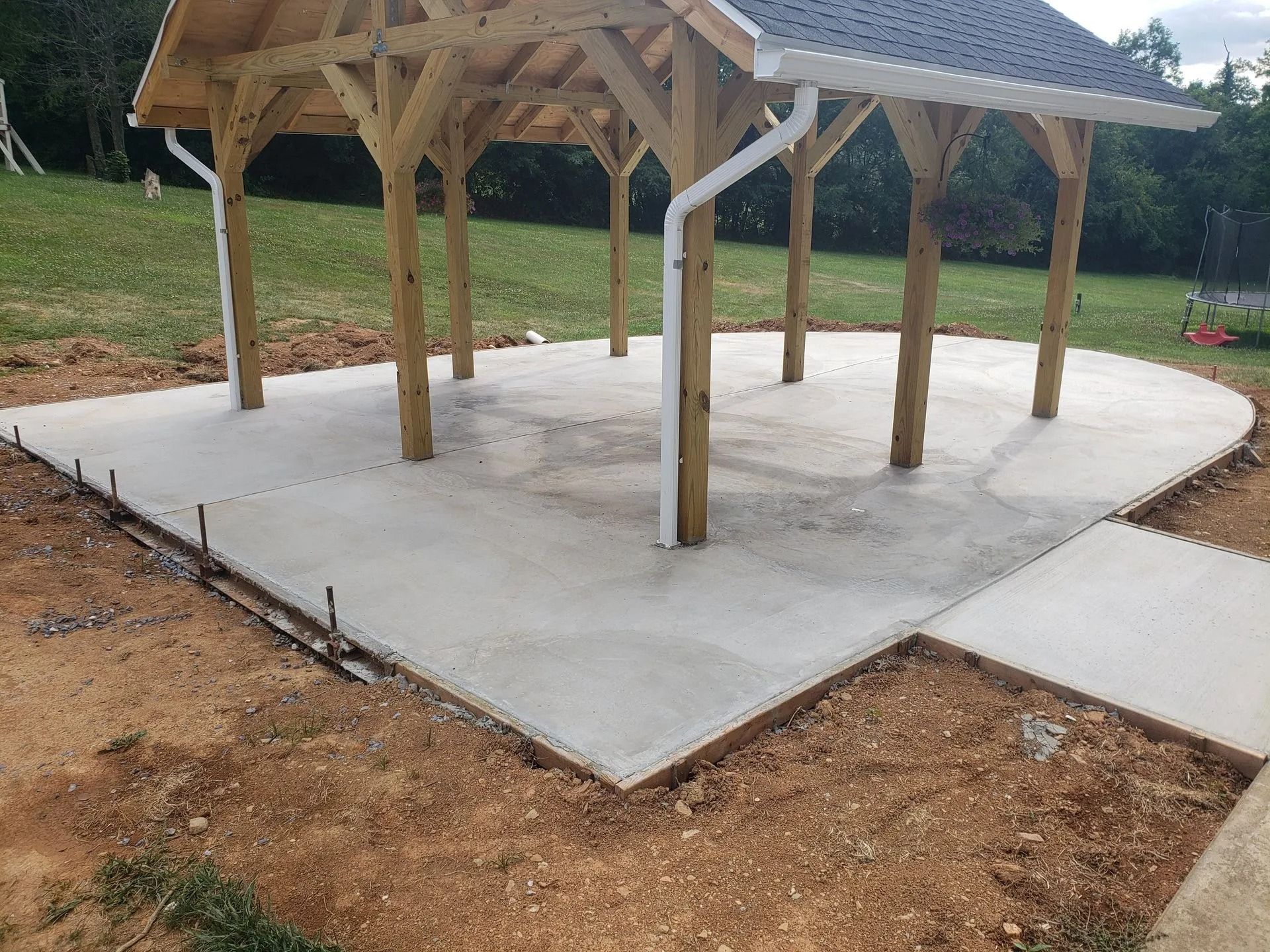 Newly poured concrete patio under a wooden gazebo roof in a grassy yard.