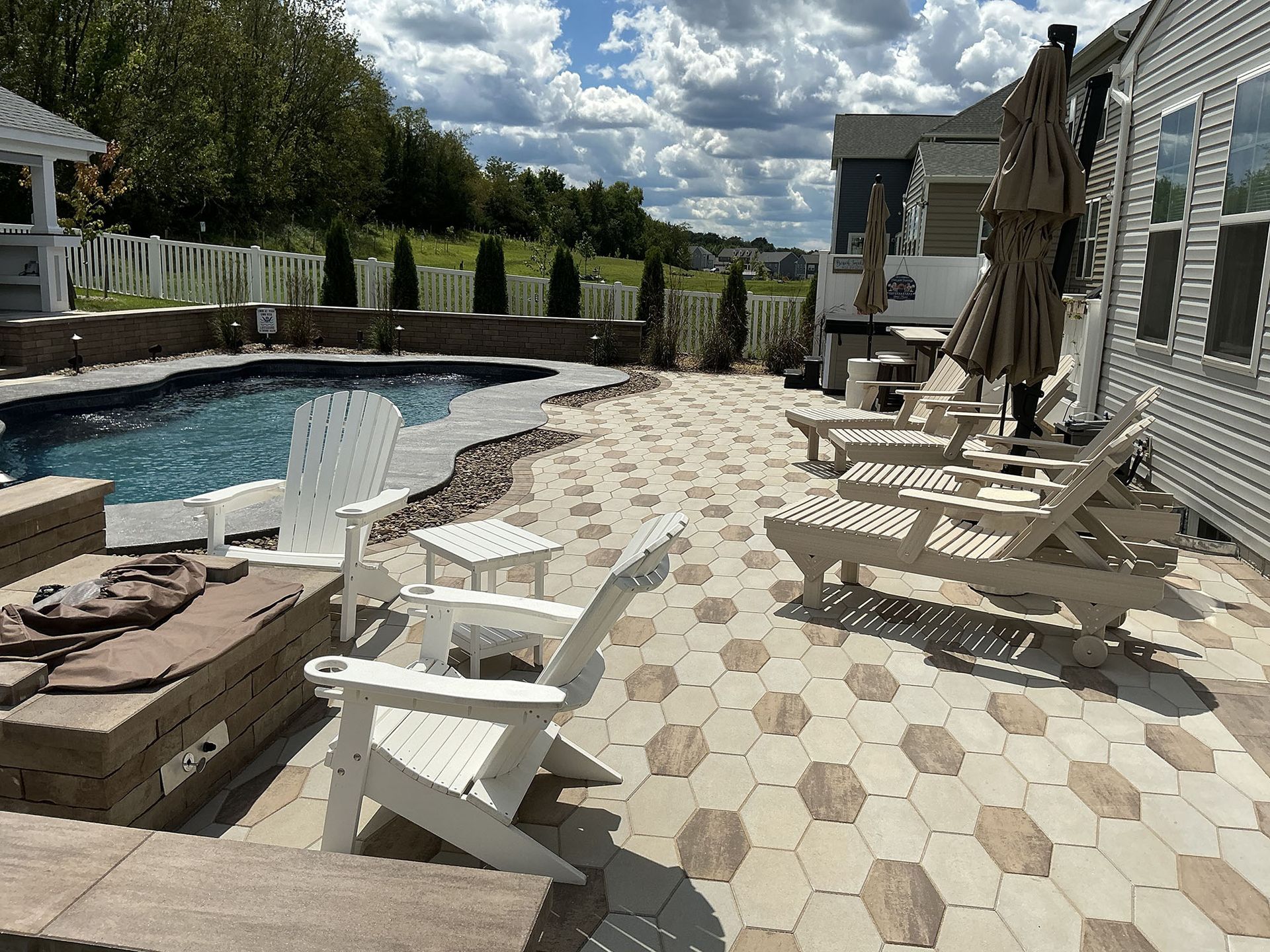 Backyard patio with pool, lounge chairs, umbrella, and patterned paving stones under a partly cloudy sky.