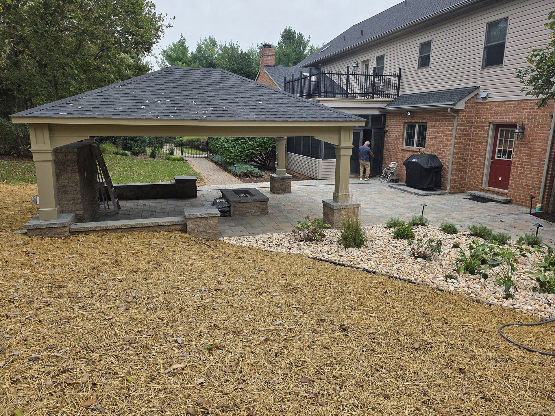 A covered outdoor patio with a seating area near a brick house and landscaped yard.
