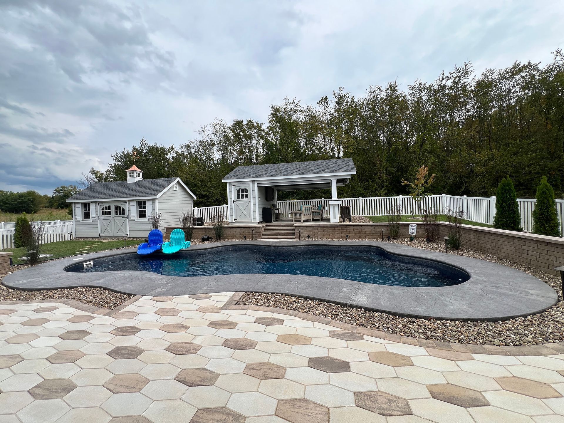 Pool area with a dark pool, patio, cabana, and a small building under an overcast sky.