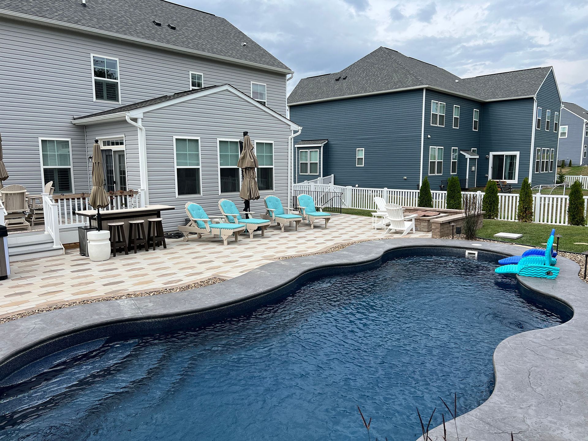 Backyard with a pool, patio, and lounge chairs. Two-story houses are in the background on a cloudy day.