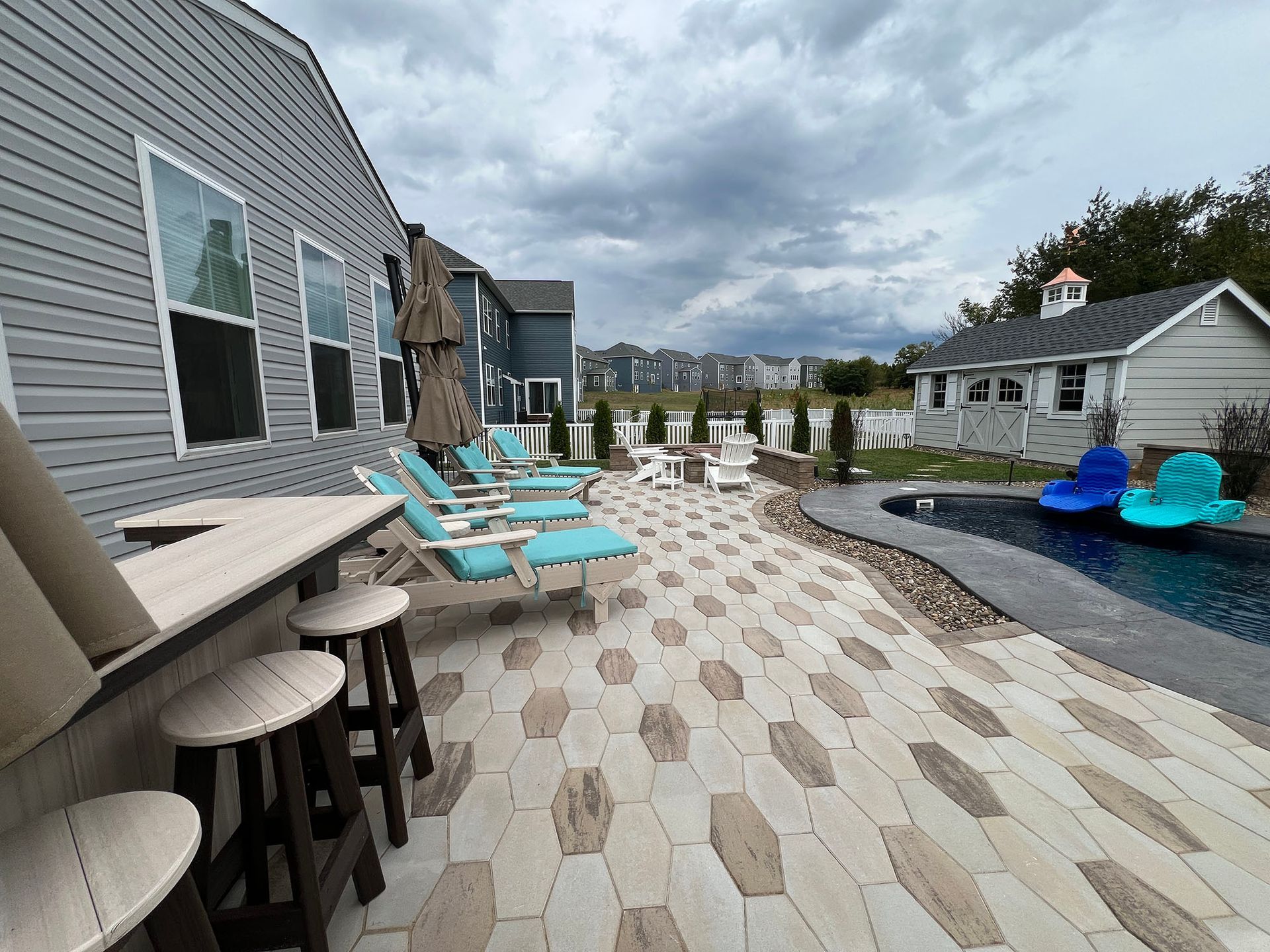Patio with pool, bar, lounge chairs, and building with gray siding, blue accents, cloudy sky.