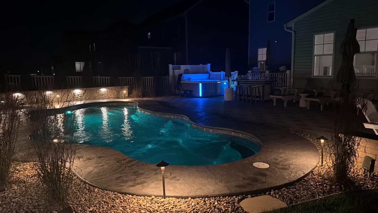 Nighttime backyard pool with blue-lit water and surrounding landscape lighting. A hot tub sits in the background.